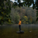 Alex standing in a yellow jacket on a lonely rock in a stream in the Belgian Ardennes on a rainy day.